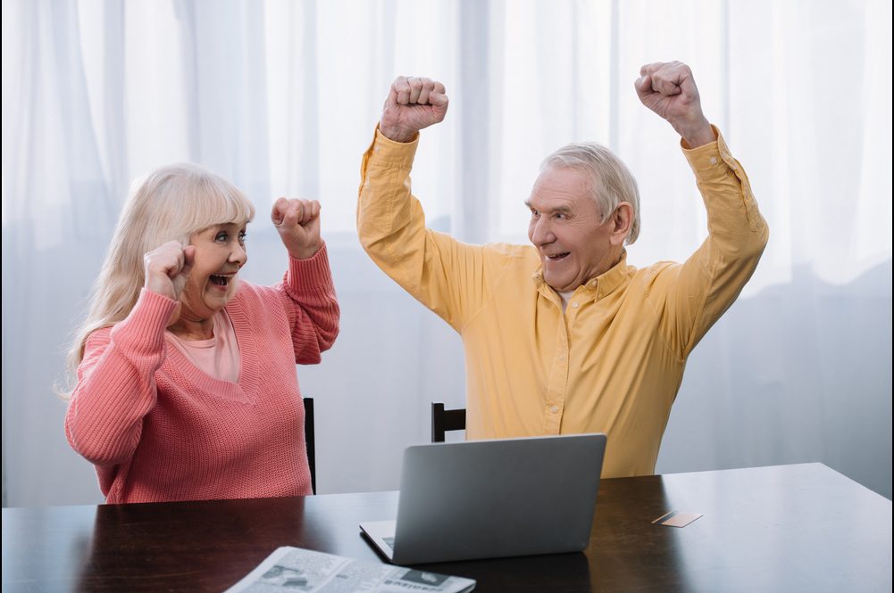 Una pareja de adultos mayores celebra con los brazos en alto frente a una computadora portátil